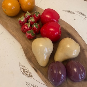 May include: A collection of ceramic fruit figurines arranged on a long wooden board. The set includes two orange oranges, six small red strawberries with green leaves, two glossy red apples, two beige pears, and two dark purple plum-shaped pieces.
