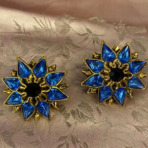 May include: A pair of gold-toned, starburst-shaped earrings. Each earring features vibrant blue, teardrop-shaped crystals surrounding a black center stone. The earrings are set against a pink, floral-patterned background.