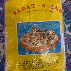 Puede incluir: Un flotador de piscina amarillo Float-A-Bar empaquetado en plástico. El embalaje presenta el texto "FLOAT-A-BAR" y "FOR ADDED FAMILY FUN - PLEASURE!". La imagen en el embalaje muestra una bandeja flotante redonda con bebidas y aperitivos.
