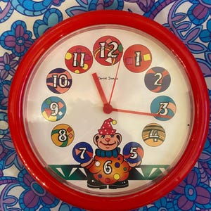 Puede incluir: Un reloj redondo con marco rojo y esfera blanca. La esfera presenta círculos numerados de colores y agujas rojas. Un personaje de oso de dibujos animados está en la parte inferior. La esfera del reloj tiene el texto "Daniel Danda".
