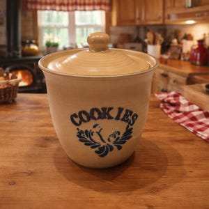 Peut inclure: Un pot à biscuits en céramique beige avec un couvercle et un bouton. Le pot porte l'inscription "COOKIES" en bleu avec un motif floral en dessous. Le pot est posé sur une surface en bois.
