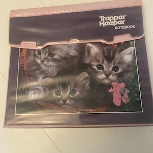 May include: A Trapper Keeper notebook with a pink clasp and a dark blue cover. The cover features a picture of three fluffy kittens in a basket with a pink flower. The text "Trapper Keeper NOTEBOOK" is printed on the top.