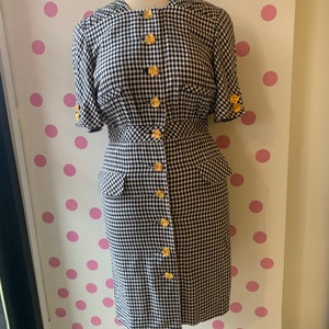 Peut inclure: Une robe en vichy noir et blanc avec des boutons dorés et des manches courtes. La robe a un corsage ajusté et une jupe légèrement évasée.