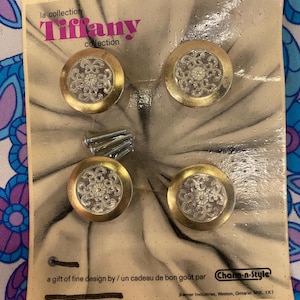 May include: Four cabinet knobs from the Tiffany collection, featuring gold-coloured rims and clear, decorative centres, presented on a light brown card. The card includes the text "Tiffany collection" and "Charm-n-Style". Screws are included.
