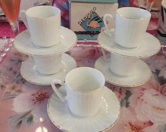 Pode incluir: Chávenas e pires de porcelana branca com acabamento dourado. As chávenas e os pires têm um design canelado. A imagem inclui uma caixa com um logótipo de flamingo. As chávenas de chá estão empilhadas numa bandeja floral rosa.