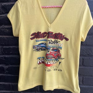 Op de afbeelding: Geel V-hals T-shirt met een afbeelding van twee klassieke auto's en de tekst "Street Masters Rod & Kustom Run-O-Dup NY 86". Het shirt hangt aan een roze en witte touwhanger.