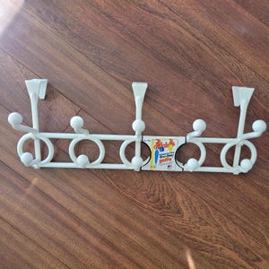 May include: A white over-the-door hook rack with a series of hooks and decorative circles. The rack has three over-the-door hooks and several smaller hooks. A small label is attached to the rack.