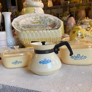 Op de afbeelding: Een verzameling vintage keukengerei, waaronder een wit-zwarte koffiepot met een zwarte handgreep en twee gele ovenschalen met blauwe bloemmotieven. Andere decoratieve items staan op de achtergrond.
