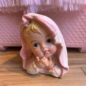 May include: A ceramic figurine of a baby girl with blonde hair, wearing a pink and white hooded towel. The baby is looking to the side and has blue eyes.
