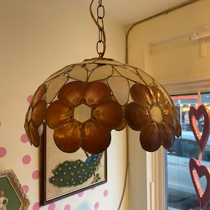 Op de afbeelding: Een witte en roze bloemvormige hanglamp met een gouden ketting. De lamp is gemaakt van schelpen en heeft een koepelvorm.