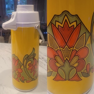 May include: A vintage yellow thermos with a white top and handle. The thermos features a floral design in shades of red, orange, and green. The thermos is cylindrical and has a pump-style dispenser.
