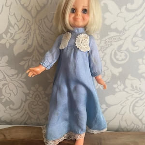 Puede incluir: Una muñeca vintage con cabello rubio y ojos azules, vestida con un vestido azul claro con ribete de encaje. El vestido de la muñeca presenta detalles de encaje en el corpiño y el dobladillo. La muñeca está de pie con los brazos extendidos.