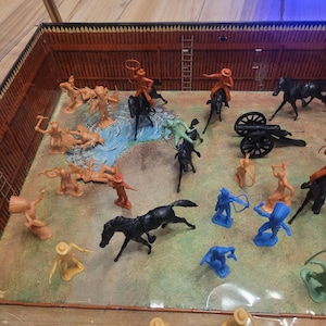 Puede incluir: Un juego de juguetes vintage que representa una escena de batalla del oeste. El juego incluye figuras de plástico en miniatura de vaqueros, nativos americanos, caballos y un cañón. Las figuras son de varios colores, incluyendo beige, azul y negro, dispuestas sobre un paisaje pintado.