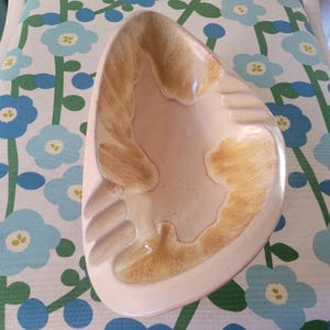 May include: A vintage ashtray in a distinctive shape, with a cream-coloured base. The ashtray has a textured, abstract design in shades of tan and brown. It has three built-in rests for cigarettes.