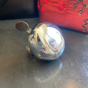 May include: A polished chrome bicycle bell with a spherical dome shape and a small metal lever attached to the side. The lever features the embossed letters WS. The reflective surface of the bell shows a distorted view of the surrounding environment.