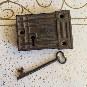 May include: A rectangular dark metal rim lock featuring a central keyhole and three circular mounting holes, accompanied by a matching skeleton key with a bow, a long cylindrical shank, and a bit at the end, both showing a weathered finish.