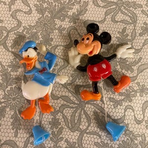 Puede incluir: Dos figuras vintage de personajes de Disney: Pato Donald con un traje de marinero azul y Mickey Mouse con pantalones cortos rojos. Ambos tienen campanas azules colgantes y están sobre un fondo estampado.