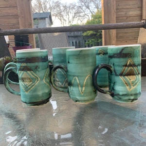 Puede incluir: Cinco tazas de cerámica con un esmalte turquesa y detalles marrones. Cada taza tiene un diseño único, que incluye formas geométricas y líneas onduladas.