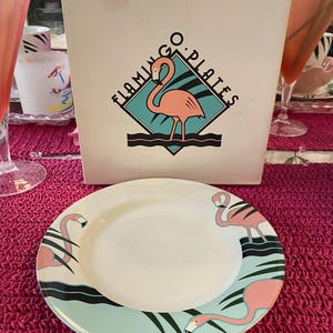 May include: A white ceramic plate with a light blue border featuring pink flamingos and black accents. The plate is part of a set, with a box that reads "Flamingo Plates."