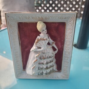 May include: A framed porcelain figurine of a woman in a white dress with gold detailing, holding a fan. The frame is white with gold trim and a burgundy background. A vintage decorative item.
