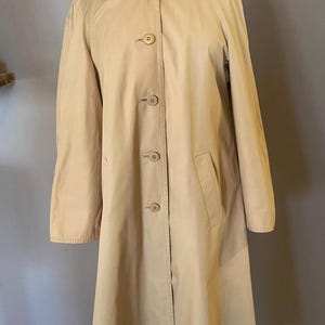 Puede incluir: Un abrigo tipo gabardina de color beige con un diseño clásico. El abrigo tiene cuello, cierre de botones y un bolsillo. El abrigo se exhibe en un maniquí.