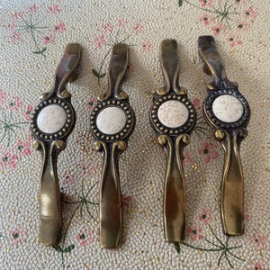 Peut inclure: Quatre poignées de tiroir en métal avec une finition en laiton antique. Chaque poignée présente une incrustation circulaire centrale en céramique blanc cassé avec de petites taches brunes, entourée d'une bordure perlée et de volutes.