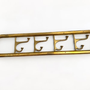 May include: A rectangular brass wall-mounted coat rack featuring a continuous outer frame and four integrated double hooks. Each hook has a curved upper and lower arm with rounded ends. The metal surface displays a weathered, aged gold finish.