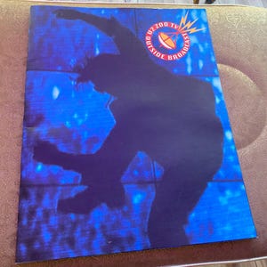 Peut inclure: Une affiche promotionnelle U2 Zoo TV Outside Broadcast. L'affiche présente une silhouette d'une personne en mouvement sur un fond bleu. Le texte "U2 Zoo TV Outside Broadcast" est visible en haut à droite.