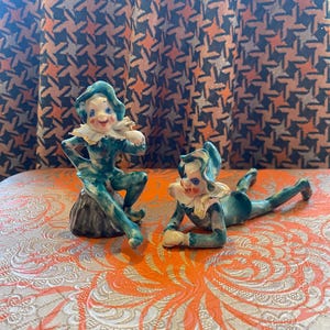 Peut inclure: Deux figurines en céramique d'elfes avec des vêtements verts et bleus, des visages blancs et des détails peints. Les elfes sont assis sur un rocher et allongés sur une surface à motifs.