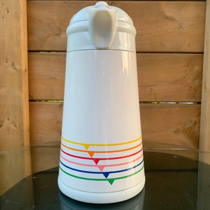 Op de afbeelding: Witte keramische kan met een kleurrijk regenboogstrepenontwerp. De strepen zijn rood, oranje, geel, groen, blauw, roze en paars. De kan heeft een handvat aan de bovenkant.