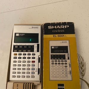 以下が含まれることがあります： ヴィンテージのSharp ELSI MATE EL-5001電子計算機とオリジナルの箱。計算機はシルバーで、黒いキーとデジタルディスプレイが付いています。箱は黄色と黒で、計算機の画像とSharpのロゴが特徴です。
