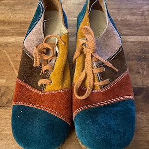 Op de afbeelding: Een paar veelkleurige suède schoenen met oranje veters. De schoenen hebben een patchwork-ontwerp met teal, bruin, oranje en gele suède panelen.