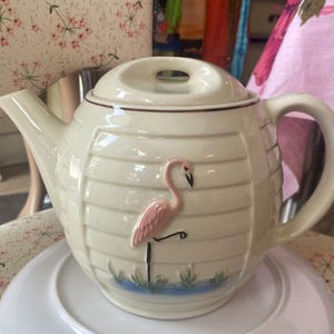 May include: A cream-coloured ceramic teapot featuring a horizontal ribbed texture and a raised pink flamingo figure standing on one leg. The design includes a small blue water detail at the base, a curved handle, a spout, and a matching lid with a handle.
