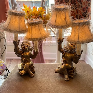 May include: Two antique table lamps with cherub bases and fringed shades. The lamps have a gold finish and are on a patterned surface. The shades are a light tan colour.