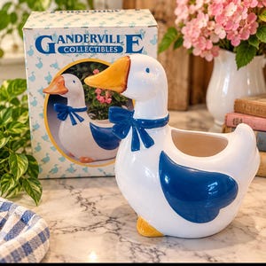 May include: A white ceramic goose planter with a glossy finish, featuring a bright orange beak, orange feet, and a dark blue bow tied around its neck. The goose has a large dark blue wing detail and an open top for plants. It sits beside its original box.