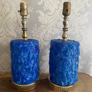 May include: A pair of vintage blue table lamps. Each lamp has a textured, cylindrical base and brass-coloured accents. The lamps feature a gold-coloured metal base and a brass-coloured neck. They are displayed on a wooden surface against a patterned wallpaper.