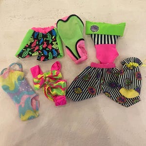 May include: A set of seven doll clothes, including a swimsuit, a skirt, a pair of shorts, a dress, a bag, and two accessories. The clothes are in a variety of colours and patterns, including pink, green, blue, black, and white.