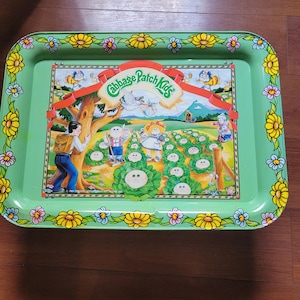 May include: A rectangular green metal serving tray featuring a Cabbage Patch Kids graphic. The border has a yellow and purple flower pattern. The center illustration depicts children in a garden with cabbage plants, a tree, and a red banner.
