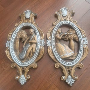 Puede incluir: Un par de placas de pared doradas y plateadas ornamentadas con figuras en relieve 3D de un hombre tocando un violonchelo y una mujer sosteniendo una flor. Las placas tienen un diseño decorativo de volutas.