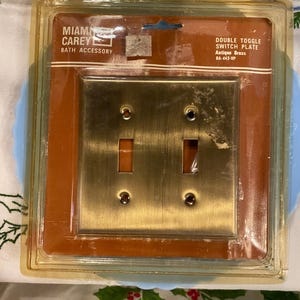 May include: A double toggle switch plate in an antique brass finish, in its original packaging. The packaging features the text "MIAMI CAREY BATH ACCESSORY" and "DOUBLE TOGGLE SWITCH PLATE Antique Brass BA-443-VP". The switch plate has two toggle openings.