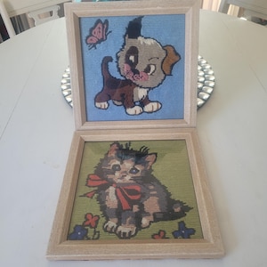 Può includere: Due quadri ricamati incorniciati. Quello superiore presenta un cucciolo con pelo marrone e marrone chiaro su sfondo blu con una farfalla rosa. Quello inferiore mostra un gattino con un fiocco rosso su sfondo verde con fiori. Entrambi sono in cornici di legno chiaro.