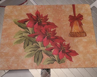 May include: Rectangular placemat with a poinsettia and bell design. The poinsettias are red with green leaves, and the bell is gold with a red bow. The background is a mottled orange color.