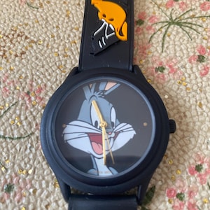 May include: A black wristwatch featuring Bugs Bunny on the watch face. The watch has a black strap with cartoon character accents. The watch hands are gold. The watch is a round shape.