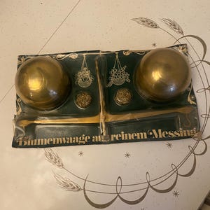 Peut inclure: Balance en laiton vintage avec deux grands bols ronds et des éléments décoratifs. La balance est montée sur une carte vert foncé avec des lettres et des illustrations dorées. La balance est probablement utilisée pour peser des articles.