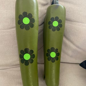 May include: A pair of olive green wooden shoe stretchers, each decorated with black and green flower motifs. They have metal handles. These stretchers are designed to preserve the shape and size of footwear.