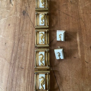 Op de afbeelding: Een armband van goudkleurig metaal met vijf witte vierkanten met gouden zeepaardjesontwerpen. De armband wordt geleverd met een bijpassend set oorbellen met hetzelfde ontwerp.