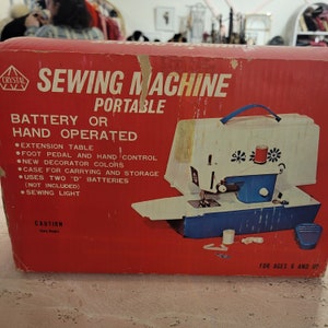 Peut inclure: Une boîte rouge et blanche avec une machine à coudre jouet bleue et blanche. La boîte indique "Sewing Machine Portable" et "Battery or Hand Operated". La boîte énumère également des fonctionnalités telles qu'une table d'extension, une pédale et un éclairage de couture. La boîte est destinée aux enfants de 6 ans et plus.