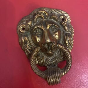 May include: A vintage brass lion head door knocker with a ring handle. The lion's face has detailed features and is mounted on a red surface. The knocker is a decorative hardware piece.