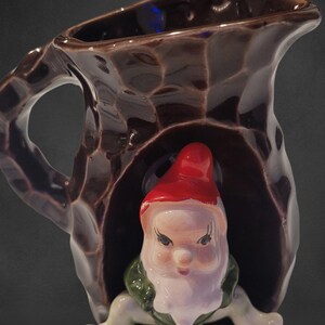 May include: A brown ceramic jug with a textured surface and a whimsical gnome figure. The gnome has a red hat, white beard, and green vest. The jug has a handle and a spout.