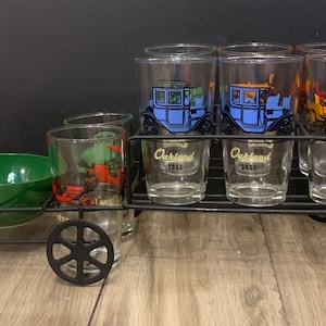 Puede incluir: Un carro de metal negro con seis vasos de vidrio y un tazón verde. Los vasos tienen imágenes de coches antiguos y el texto "Oakland 1911" y "Ford 1909".